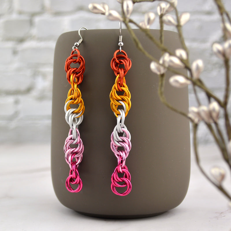 Long and thin chainmaille spiral earrings hang from a grey tumbler against a light grey brick wall and light grey marble surface. Earrings are the color of the lesbian pride flag: dark orange, bright orange, white, pink and fuchsia Earrings resemble a strand of DNA.