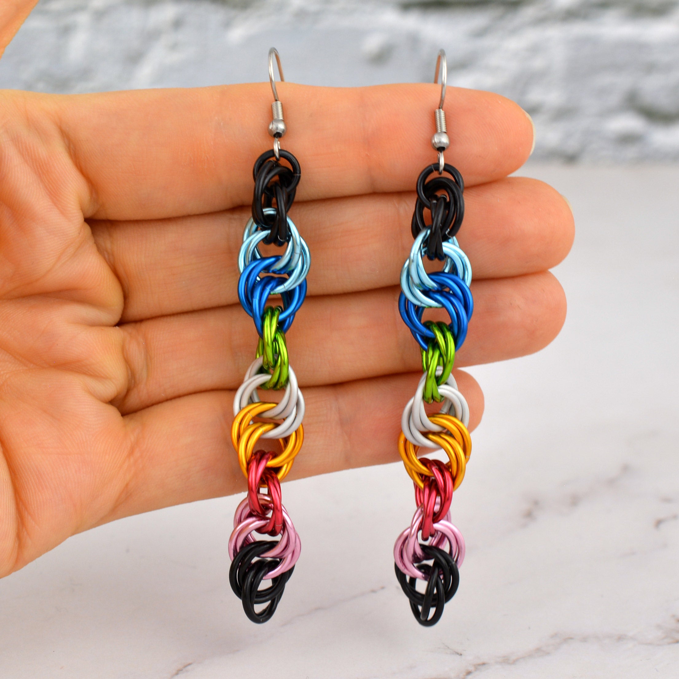 Artisan Rebeca Mojica holds long, spiral earrings in hand. The black, light blue, blue, bright green, white, gold, dark pink, light pink aluminum rings form a helix pattern representing the queer pride flag. A soft white and grey blurred background.