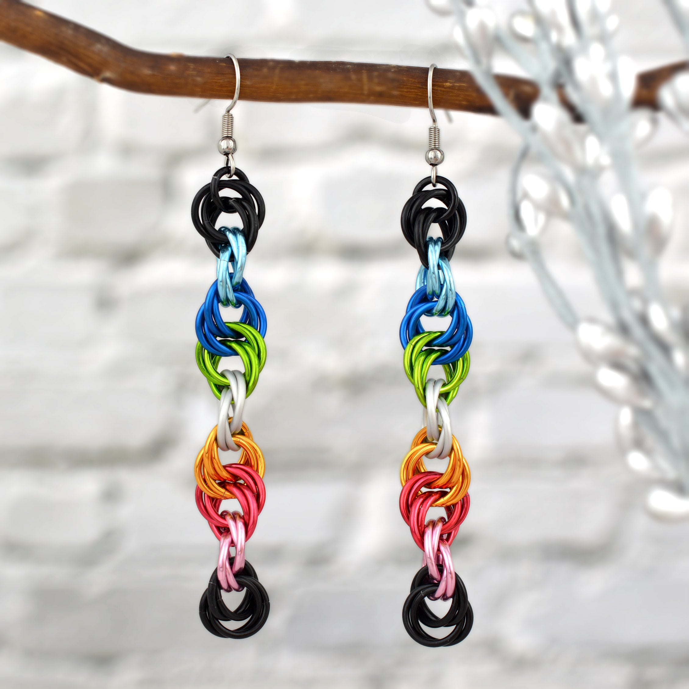 Long, colorful spiral earrings crafted with anodized aluminum jump rings (black, light blue, blue, bright green, white, gold, dark pink, light pink). Silver hooks hang them from a thin brown branch against a blurred white brick wall.