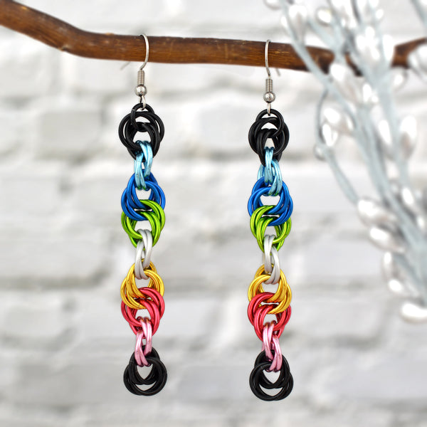 Long, colorful spiral earrings crafted with anodized aluminum jump rings (black, light blue, blue, bright green, white, gold, dark pink, light pink). Silver hooks hang them from a thin brown branch against a blurred white brick wall.