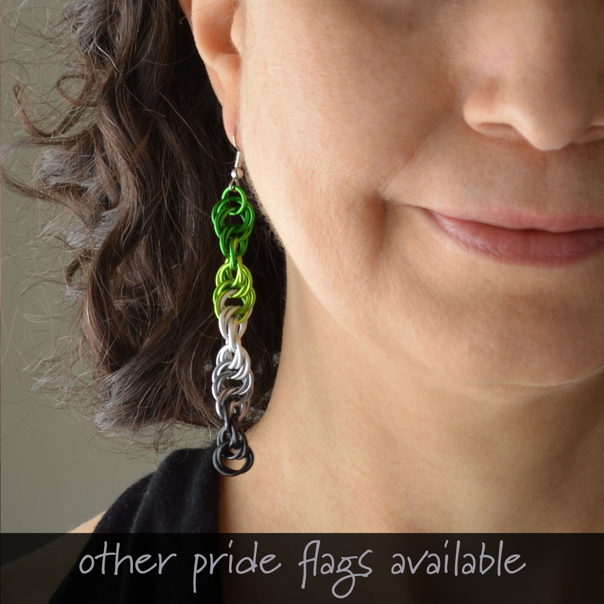 Close-up of Rebeca Mojica modeling a long aromantic pride earring in green, light green, white, grey, and black chainmaille. Stainless steel hook. "other pride flags available" text shown.