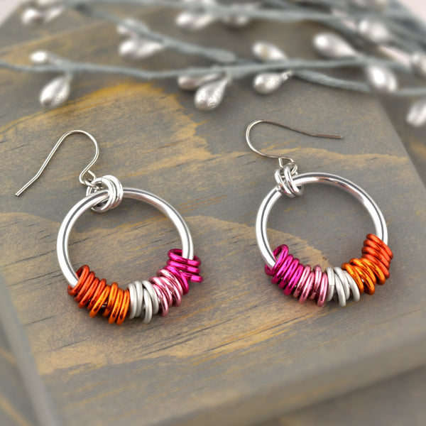 Lesbian pride earrings displayed on grey wood. The chainmaille earrings by Rebeca Mojica have a large silver color hoop, and on the silver hoop are 20 tiny links in the colors of the lesbian pride flag (4 links in each color: two shades of pink, two shades of orange + white). The tiny colors are mirror image on the second earring.