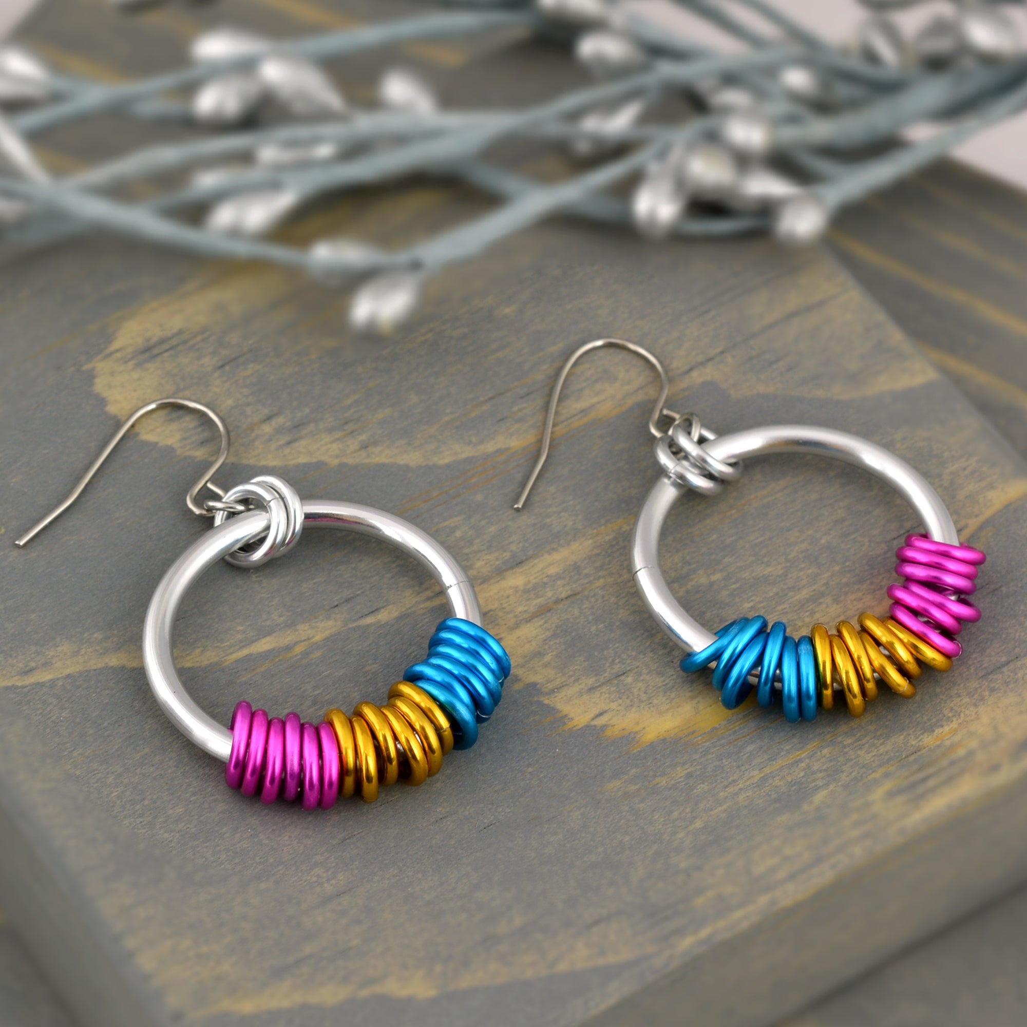 Pansexual pride chainmaille earrings by Rebeca Mojica displayed on grey wood. Earrings have a large silver color hoop, and on the silver hoop are 18 tiny links; six links in each of the 3 colors of the pan pride flag (pink, gold and azure)