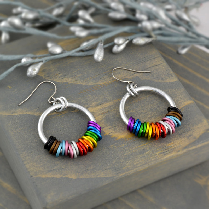 LGBTQ pride chainmaille hoop earrings by Rebeca Mojica on grey wood. Each earring has a large hoop link with 22 tiny links; two links in each of the 11 colors of the rainbow progress pride flag