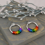 Rainbow pride hoop earrings handmade by Rebeca Mojica on grey wood. Earrings have a large silver color hoop, and on the silver hoop are 18 tiny links; three links in each of the 6 colors of the rainbow pride flag