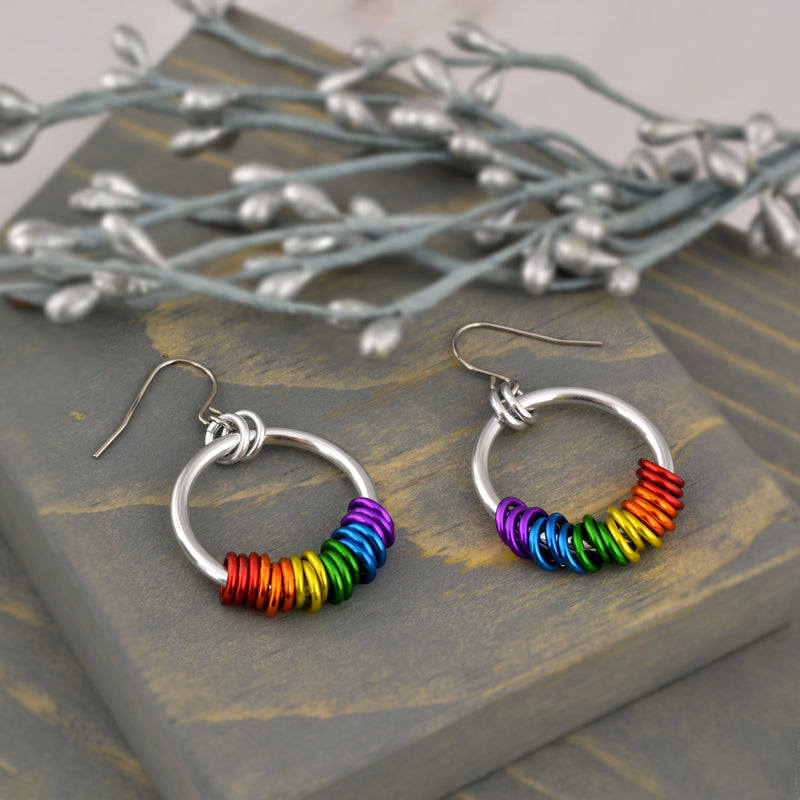 Rainbow pride hoop earrings handmade by Rebeca Mojica on grey wood. Earrings have a large silver color hoop, and on the silver hoop are 18 tiny links; three links in each of the 6 colors of the rainbow pride flag