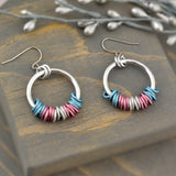 Transgender pride chainmaille hoop earrings by Rebeca Mojica displayed on grey wood. Earrings have a large silver color hoop, and on the silver hoop are 20 tiny links; four links in each of the 5 stripes of the trans pride flag (blue, pink and white).