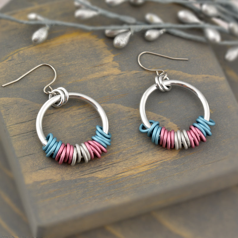 Transgender pride chainmaille hoop earrings by Rebeca Mojica displayed on grey wood. Earrings have a large silver color hoop, and on the silver hoop are 20 tiny links; four links in each of the 5 stripes of the trans pride flag (blue, pink and white).