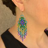 Closeup of lng statement chainmaille earring in peacock colors being worn by the artisan, Rebeca Mojica. Earrings are 4" long and have links in teal, purple, lime, blue and violet.