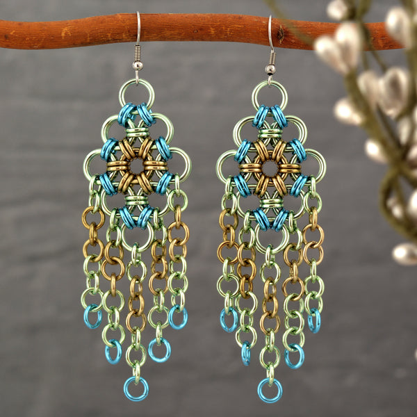 A pair of intricate chainmaille chandelier earrings hanging from a small tree branch with a blurred grey background. Earrings are pastel green, beige and light blue