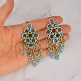 Artisan Rebeca Mojica holds a pair of Seashell colorway chainmaille earrings at a distance to show their full length.