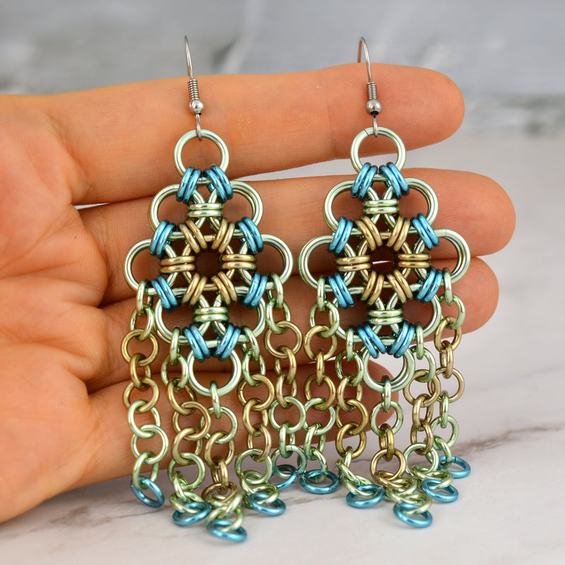 Close-up of artisan Rebeca Mojica holding a pair of Seashell colorway chainmaille earrings, highlighting the intricate chainmaille weave and colors: pastel mint green, beige and light blue