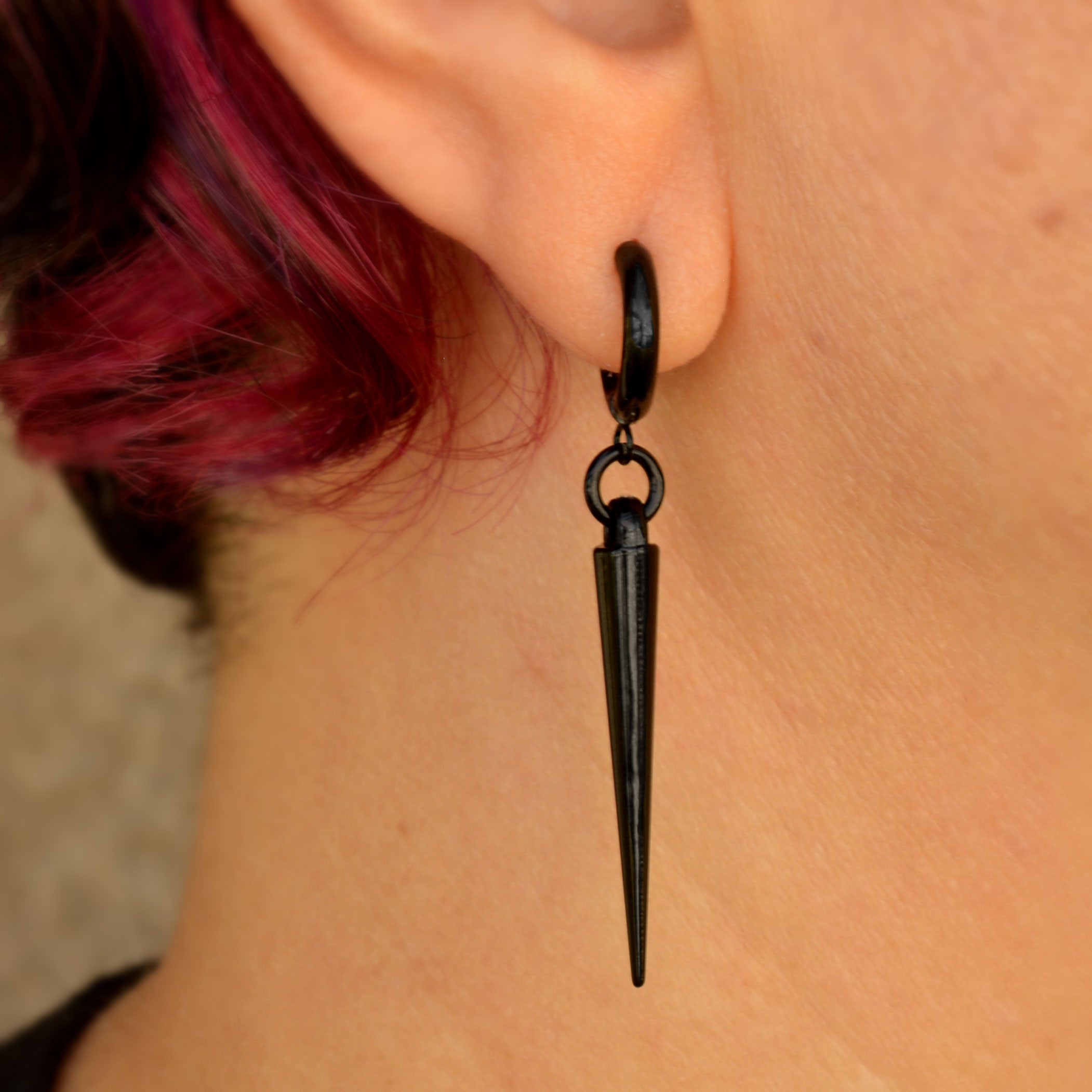 Closeup of a long black spike hoop huggie earring being worn. There is a small black ring connecting the shiny black huggie to an acrylic spike. Artisan Rebeca Mojica is wearing the earring; her short red and black hair is visible at left