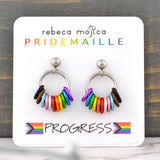From each ball hangs a steel hoop with 11 hanging rings in the colors of the progress pride flag: black, brown, light blue, light pink, white, red, orange, yellow, green, blue, violet.. The earring card reads Rebeca Mojica at top, the second line reads PRIDEMAILLE in rainbow color fonts. The bottom of the card has a sticker with the word PROGRESS flanked by 2 progress pride flags