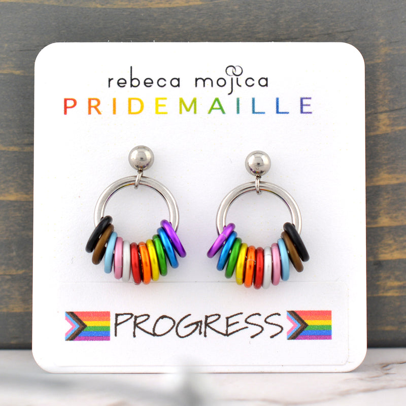 From each ball hangs a steel hoop with 11 hanging rings in the colors of the progress pride flag: black, brown, light blue, light pink, white, red, orange, yellow, green, blue, violet.. The earring card reads Rebeca Mojica at top, the second line reads PRIDEMAILLE in rainbow color fonts. The bottom of the card has a sticker with the word PROGRESS flanked by 2 progress pride flags