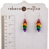 A pair of Rebeca Mojica Jewelry's Mini LGBTQ Pride Leaf Dangle earrings, featuring tiny colorful aluminum leaves, displayed on a branded measuring board next to a ruler, showing their approximate length.
