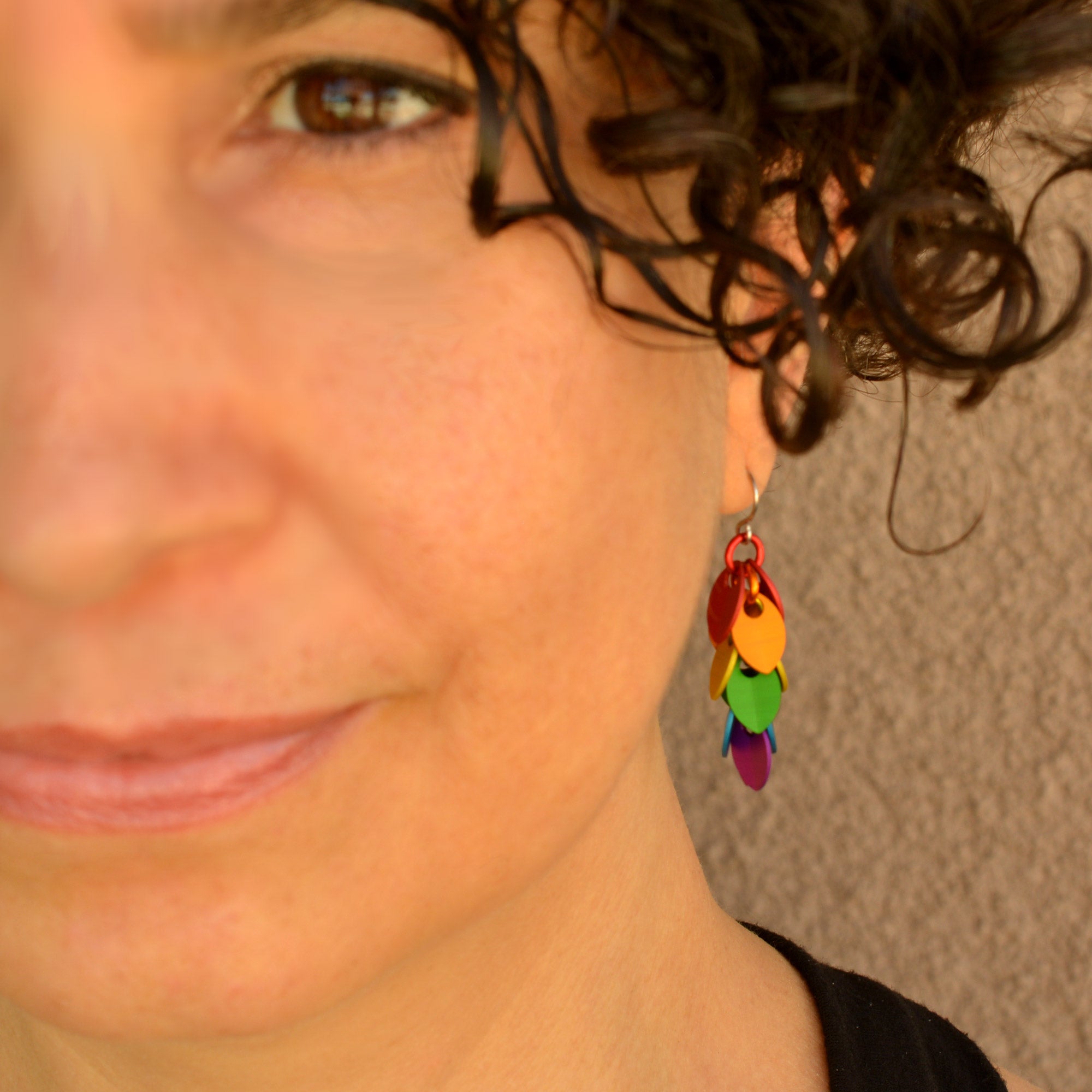 Close-up of Rebeca Mojica, a person with curly dark hair and a warm smile, wearing a "mini" rainbow cascading leaves earring from Rebeca Mojica Jewelry.