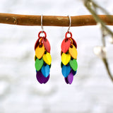 A pair of mini cascading leaves LGBTQ rainbow pride earrings by Rebeca Mojica Jewelry, featuring colorful aluminum leaves (scales), hanging from a natural tree branch with blurred white brick wall in the background.