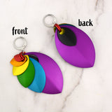 An aerial view of two rainbow stacked leaf earrings on a marble surface. The earring on the left shows the 'front' with the vibrant nested leaves, splayed out. The earring on the right shows the 'back' with a single, solid violet leaf and a small black scale visible.