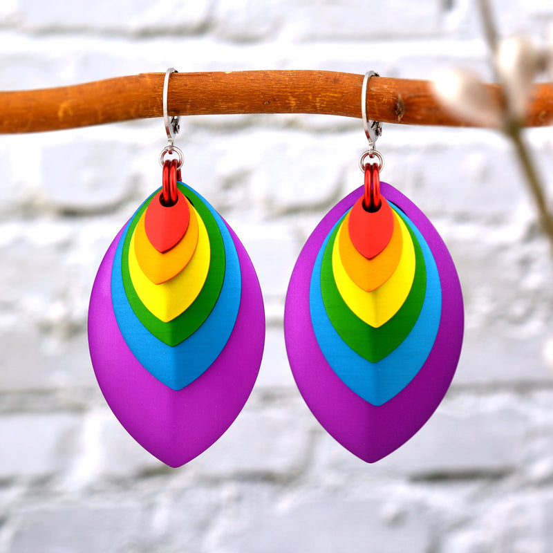  pair of rainbow-colored stacked leaf earrings, featuring red, orange, yellow, green, blue, and purple leaves, hanging from a natural brown tree branch. A blurred white brick wall is in the background.
