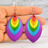 A close-up of artisan Rebeca Mojica holding a pair of vibrant rainbow-colored stacked leaf earrings in her fingertips. The earrings feature nested aluminum leaves in red, orange, yellow, green, azure and violet