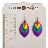 A pair of rainbow-colored stacked leaf earrings with stainless steel fishhook ear wires, placed on a wooden display board with a ruler. The ruler shows the earrings are approximately 2.75 inches long, or about 7 centimeters. The brand name 'rebeca mojica' is visible at the top of the board.