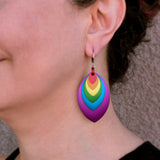A close-up of artisan Rebeca Mojica’s left ear, showing a vibrant rainbow-colored stacked leaf earring being worn. The bottom of the earring hangs about midway between the earlobe and the shirt line .Rebeca has dark, curly hair and is wearing a black top