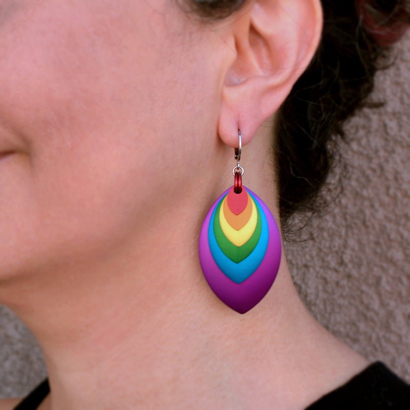 A close-up of artisan Rebeca Mojica’s left ear, showing a vibrant rainbow-colored stacked leaf earring being worn. The bottom of the earring hangs about midway between the earlobe and the shirt line .Rebeca has dark, curly hair and is wearing a black top