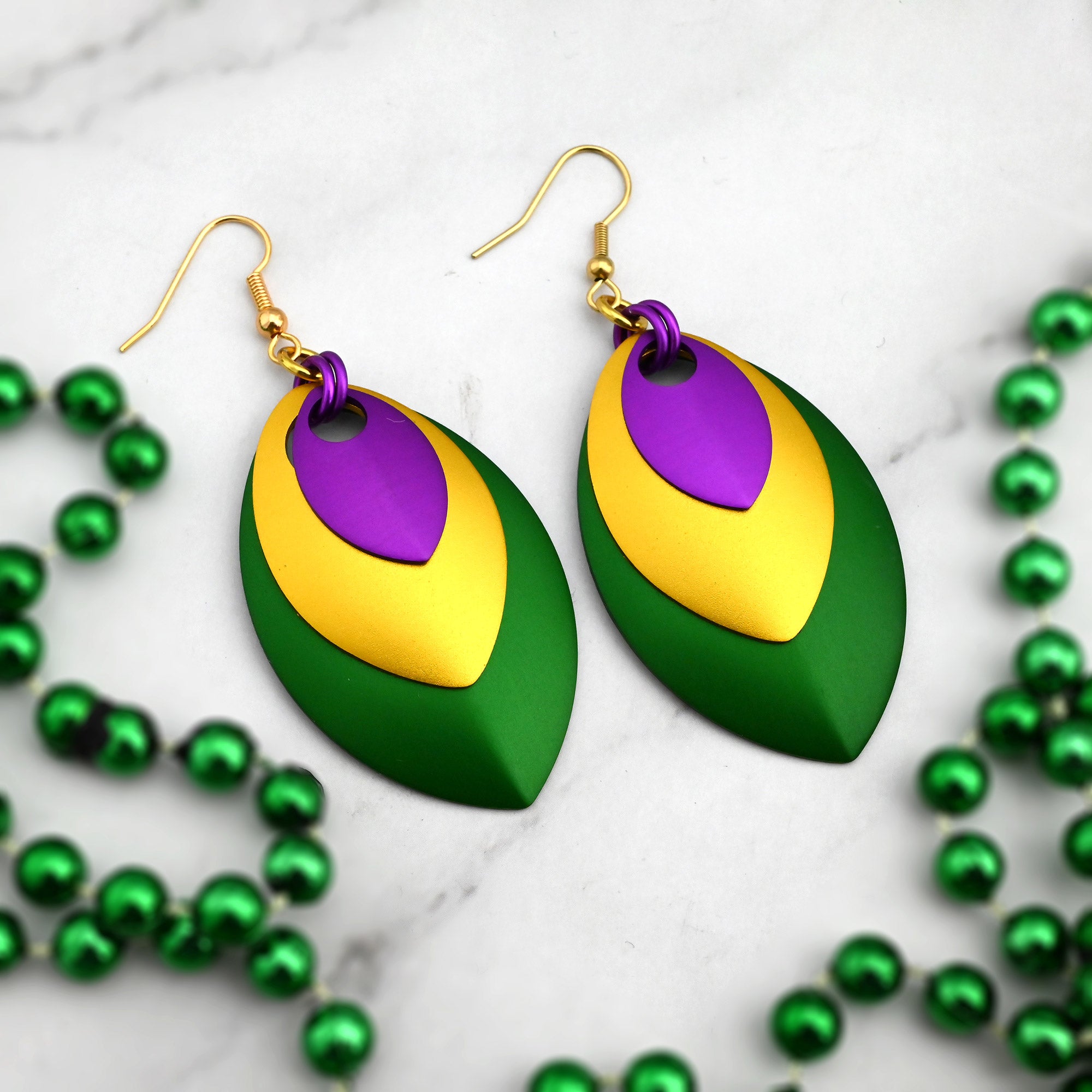 Flat lay of bold nested leaf earrings in Mardi Gras colors on white marble surrounded by metallic green festive beads.