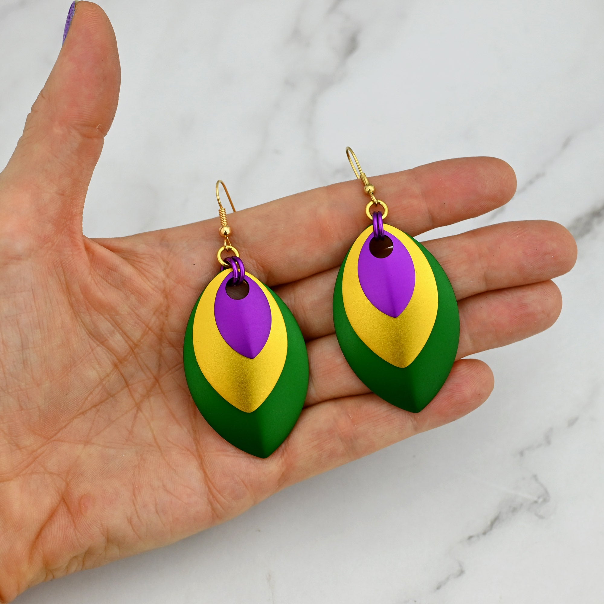 A pair of large stacked leaf Mardi Gras earrings in vibrant violet, gold, and green colors. The earrings are resting in a palm to show their scale; they span nearly the entire width of all four fingers.