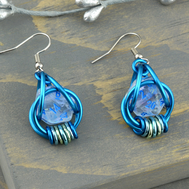 Closeup of chainmaille dice earrings by Rebeca Mojica on grey wood. Each earring has a mini translucent 20-sided die with blue numbers, encased in a 4-ring blue aluminum cage, with 7 tiny rings hanging from the bottom in shades of blue and pastel green. 