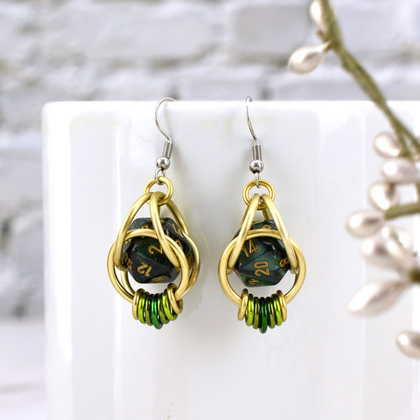 Closeup of chainmaille dice earrings by Rebeca Mojica hanging on a white mug. Each earring has a mini dark green marbled 20-sided die with gold numbers, encased in a 4-ring light gold color aluminum cage, with 7 tiny rings hanging from the bottom in an ombre from gold to green and back to gold. 