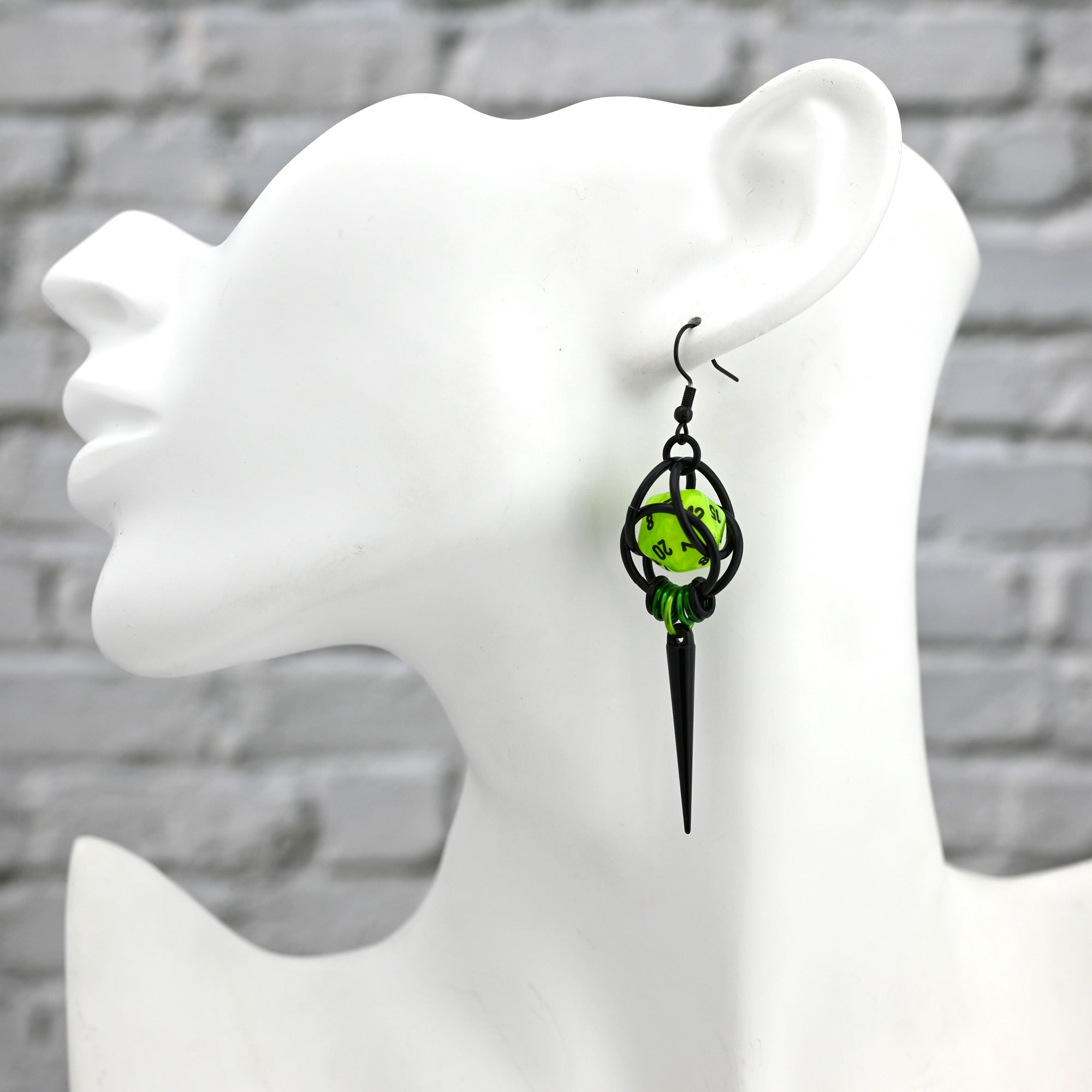 A single Neon Toxic Green D20 spike earring shown on a white mannequin head to demonstrate scale and drape. The chartreuse miniature die is held in a black chainmaille cage, hanging roughly 3.25 inches long against a white-washed brick background.