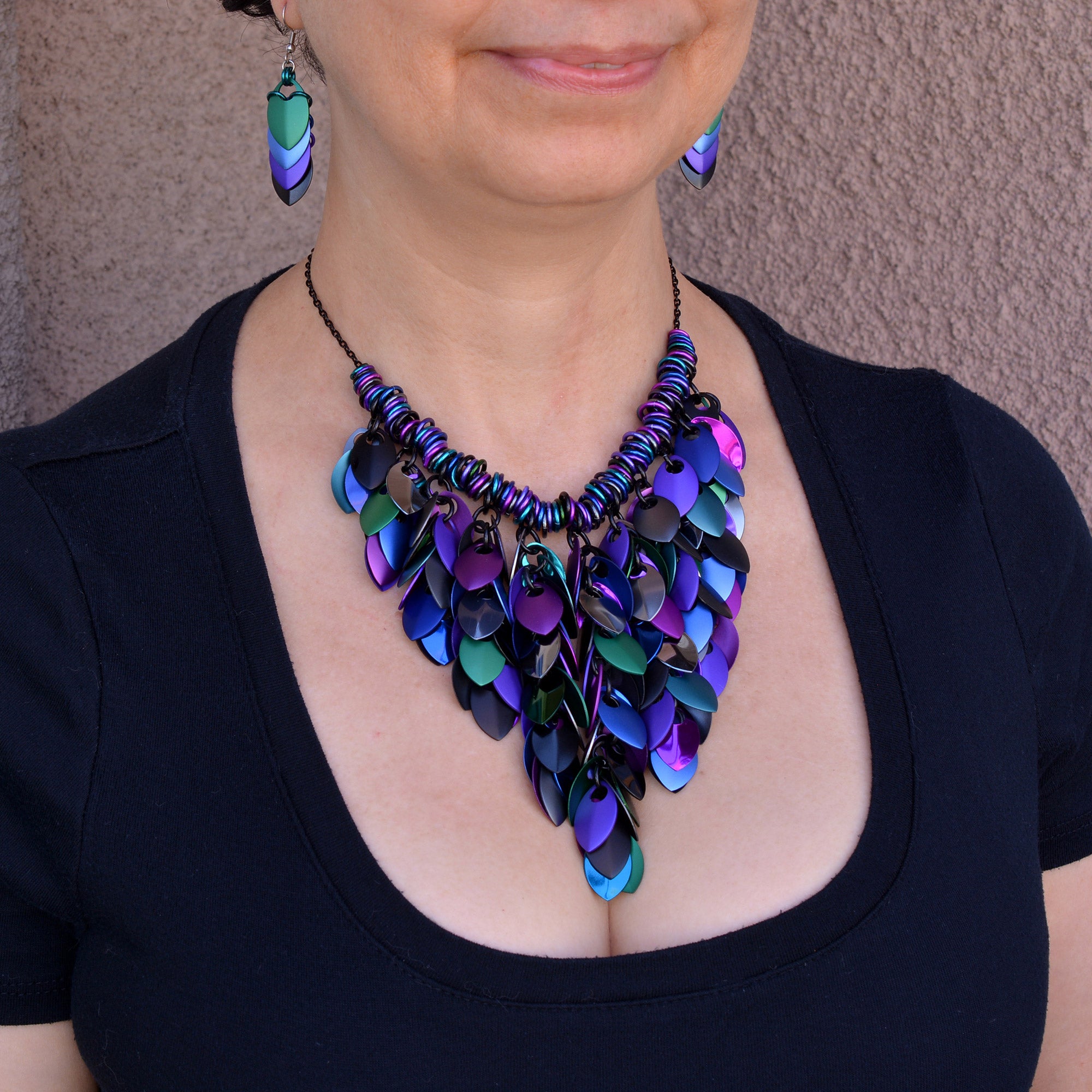 Artisan Rebeca Mojica wearing her statement scalemaille bib necklace in Dark Mermaid colors, featuring purple, blue, black and grey anodized aluminum scales ("leaves").