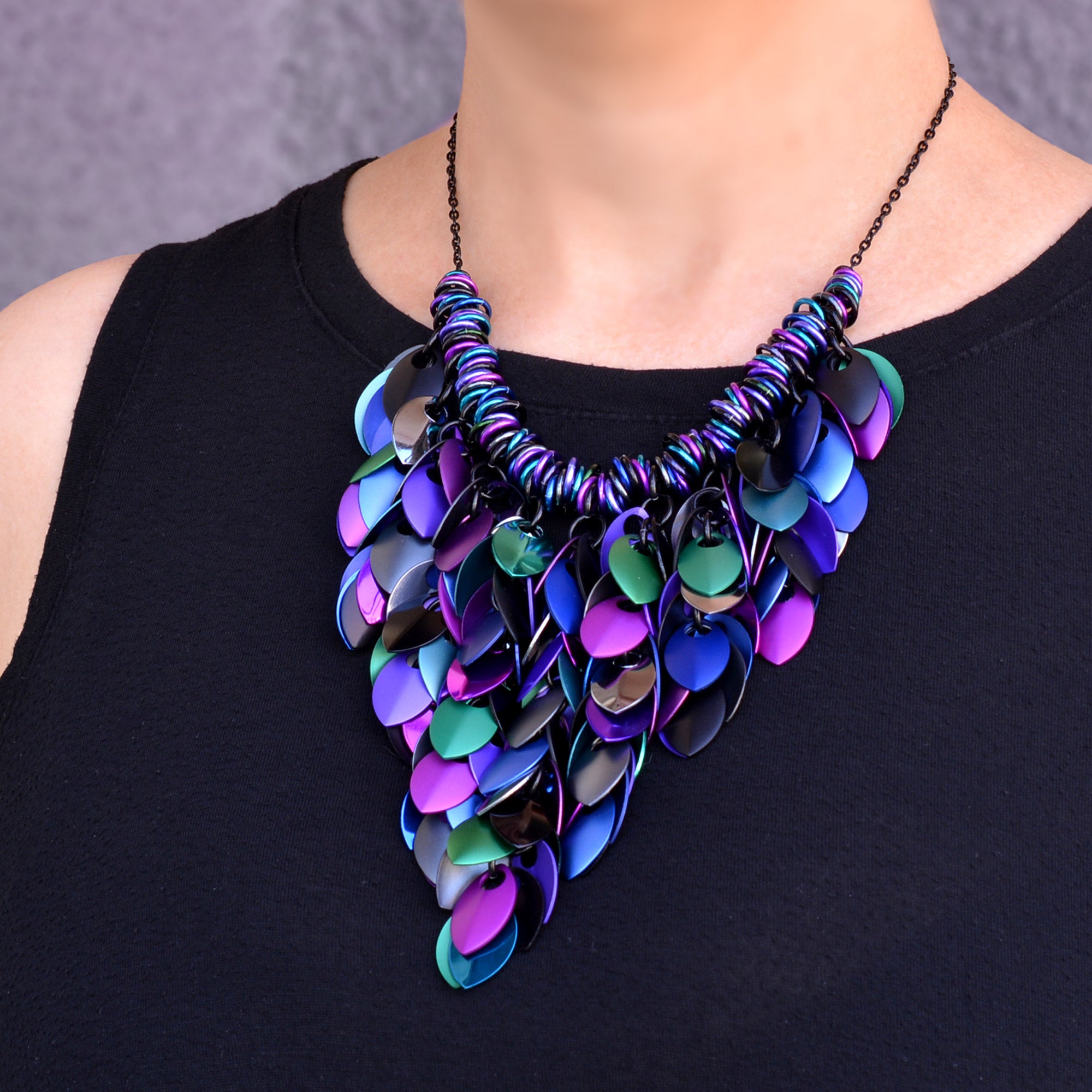 A close-up view of the Feathered Bib necklace in "Dark Mermaid" worn against a black top. The shot highlights the intricate layering of over 150 metal scales in iridescent shades of emerald green, royal blue, deep purple, and black. The scales are arranged in a sleek, downward-tapering V-shape, hanging from a multi-color of jump rings arranged in a coil.