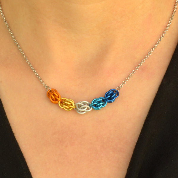 Closeup of short aroace pride chainmaille necklace being worn. Necklace has a 2-inch focal section attached to a thin steel chain on each end. The focal is made of 5 chainmaille “beads” in the Sweet Pea weave - each segment has 6 links of the same color. From left to right, colors are bright orange, gold, white, bright blue and dark blue - to best match the aromantic asexual pride flag.