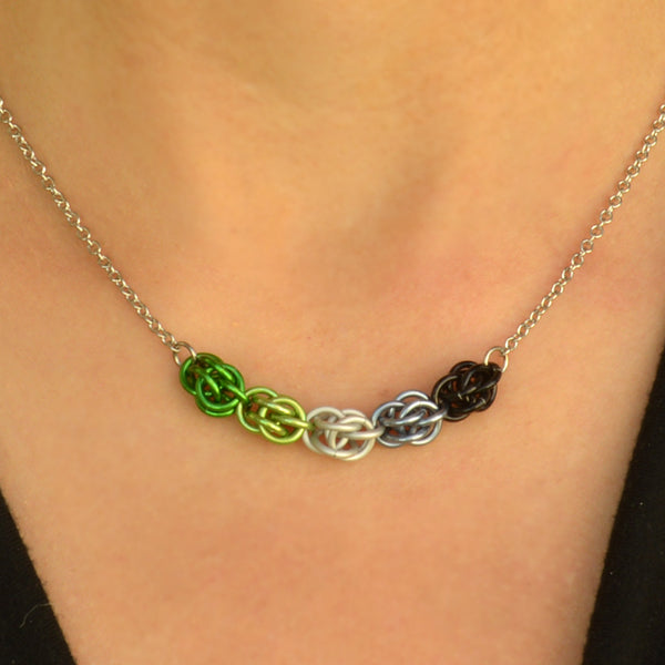 Closeup of short chainmaille necklace being worn. Necklace has a 2-inch focal section attached to a thin steel chain on each end. The focal is made of 5 chainmaille “beads” in the Sweet Pea weave - each segment has 6 links of the same color. From left to right, colors are green, lime, white, grey and black - to best match the aromantic pride flag.