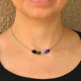 Minimalist asexual pride chainmaille necklace being worn. Necklace has a 2-inch focal section attached to a thin steel chain on each end. The focal is made of 4 chainmaille “beads” in the Sweet Pea weave - each segment has 6 links of the same color. From left to right, colors are bright purple, white, grey and black - to best match the asexual pride flag