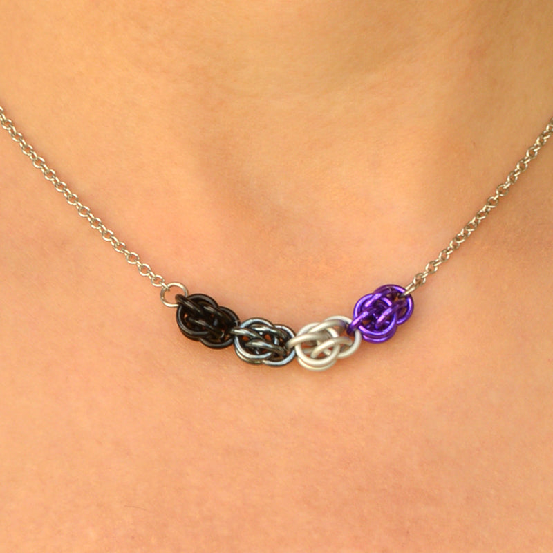 Closeup of short ace pride chainmaille necklace being worn. Necklace has a 2-inch focal section attached to a thin steel chain on each end. The focal is made of 4 chainmaille “beads” in the Sweet Pea weave - each segment has 6 links of the same color. From left to right, colors are bright purple, white, grey and black - to best match the asexual pride flag
