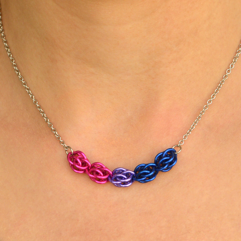 Closeup of short bi pride chainmaille necklace being worn. Necklace has a 2-inch focal section attached to a thin steel chain on each end. The focal is made of 5 chainmaille “beads” in the Sweet Pea weave - each segment has 6 links of the same color. From left to right, colors are pink, lilac and blue- to best match the bisexual pride flag.