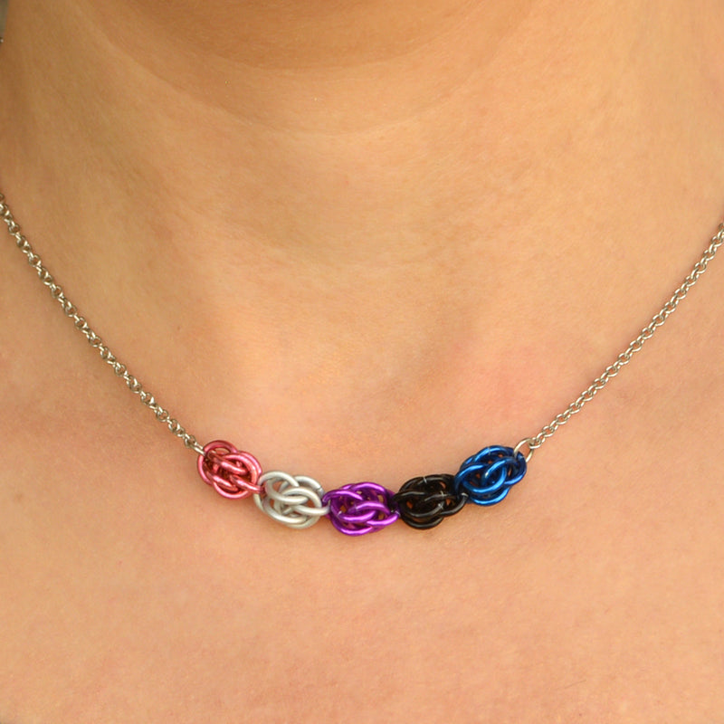 Closeup of short chainmaille necklace being worn. Necklace has a 2-inch focal section attached to a thin steel chain on each end. The focal is made of 5 chainmaille “beads” in the Sweet Pea weave - each segment has 6 links of the same color. From left to right, colors are pink, white, purple, black and blue - to best match the genderfluid pride flag.