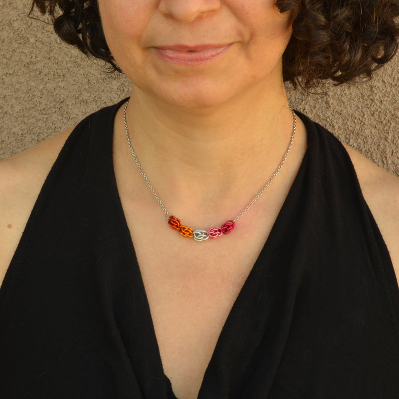 lesbian pride necklace worn on a smiling woman with a plunging V-neck black top. The necklace is minimalist, in the colors of the lesbian pride flag