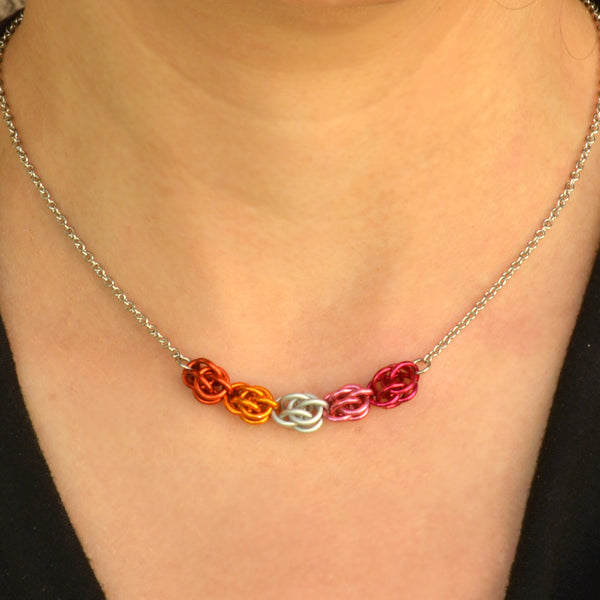 Closeup of short chainmaille necklace being worn. Necklace has a 2-inch focal section attached to a thin steel chain on each end. The focal is made of 5 chainmaille “beads” in the Sweet Pea weave - each segment has 6 links of the same color. From left to right, colors are papaya, orange, white, pink and fuchsia - to best match the lesbian pride flag.