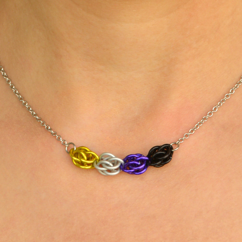 Closeup of short nonbinary pride chainmaille necklace being worn. Necklace has a 2-inch focal section attached to a thin steel chain on each end. The focal is made of 4 chainmaille “beads” in the Sweet Pea weave - each segment has 6 links of the same color. From left to right, colors are yellow, white, purple and black - to best match the nonbinary pride flag.