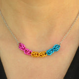 Closeup of short chainmaille pan pride necklace being worn. Necklace has a 2-inch focal section attached to a thin steel chain on each end. The focal is made of 6 chainmaille “beads” in the Sweet Pea weave - each segment has 6 links of the same color. From left to right, colors are pink, gold and blue - to best match the pansexual pride flag.