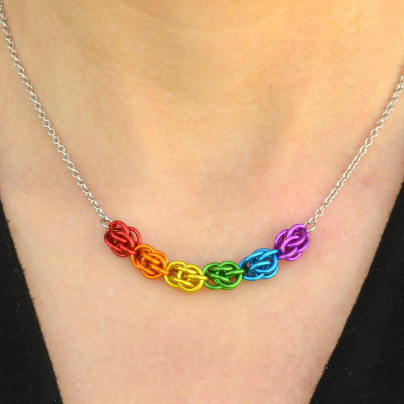 Closeup of short chainmaille necklace being worn. Necklace has a 2-inch focal section attached to a thin steel chain on each end. The focal is made of 6 chainmaille “beads,” each with 6 links of the same color. From left to right, colors are red, orange, yellow, green, blue and purple.