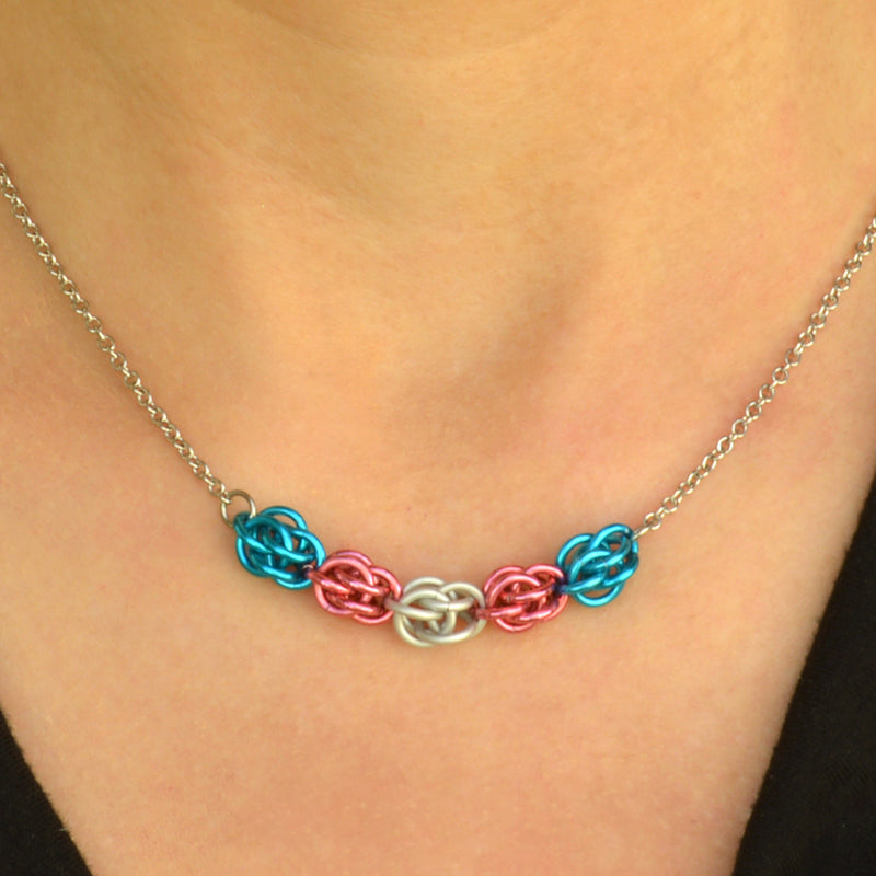 Closeup of short transgender pride chainmaille necklace being worn. Necklace has a 2-inch focal section attached to a thin steel chain on each end. The focal is made of 5 chainmaille “beads” in the Sweet Pea weave - each segment has 6 links of the same color. From left to right, colors are bright blue, pink, white, pink and bright blue - to best match the trans pride flag.