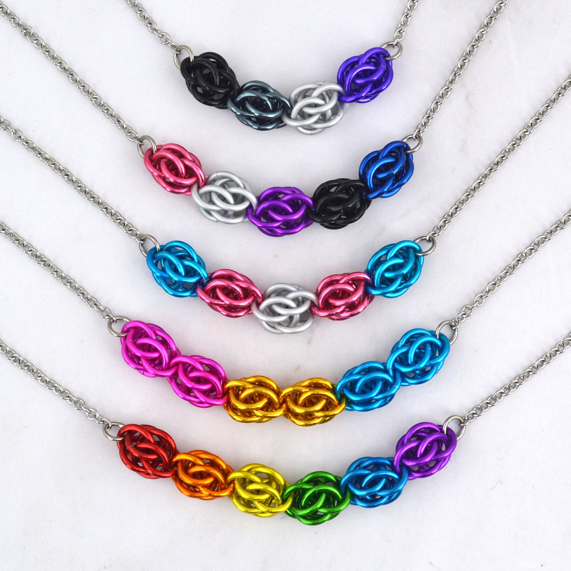 The center pieces of five colorful chainmaille necklaces arranged from shortest to longest. The colors in each necklace represent LGBTQ pride flags - from top to bottom: asexual, genderfluid, transgender, pansexual and rainbow