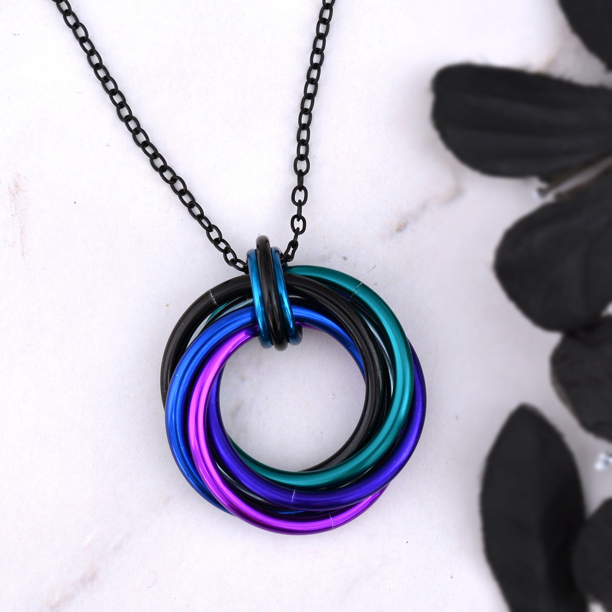 Swirl Pendant necklace in Dark Mermaid colors on a black chain, photographed on a white marble surface next to a black floral accent. The 6 large links are black, grey, teal, purple, violet and blue.