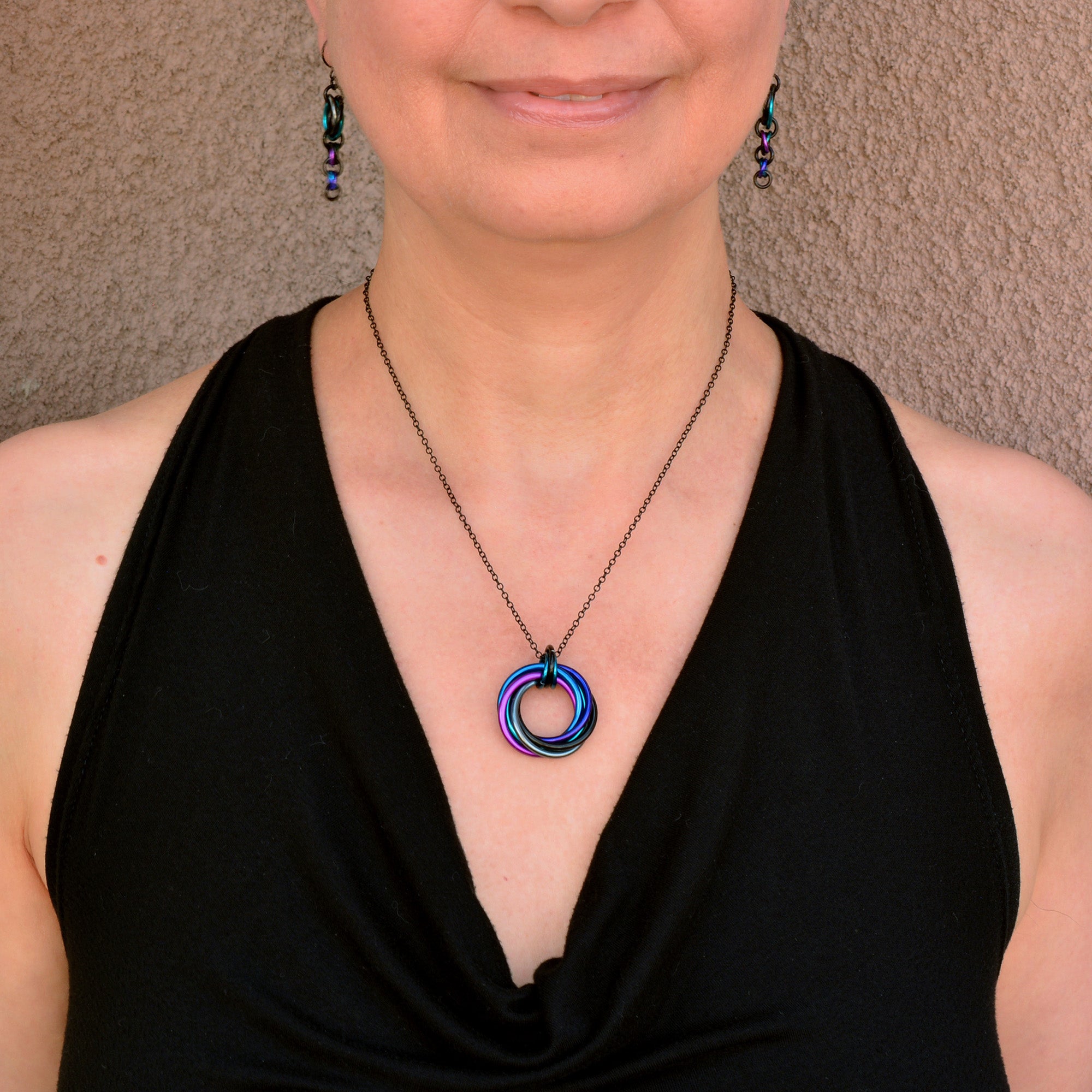 Artisan Rebeca Mojica models the Swirl Pendant necklace in Dark Mermaid (black, grey, teal, purple, violet, blue) and matching Comet Earrings.